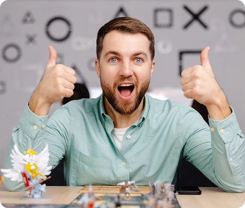 Een man met een baard in een lichtgroen shirt glimlacht breed en steekt beide duimen omhoog. Hij zit aan een tafel met spelstukken. De sfeer is vrolijk.